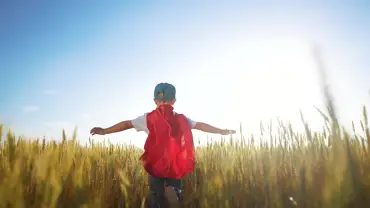 Young boy running through a wheat field wearing a baseball cap, jeans, white shirt, and superhero cape with arms outstretched.