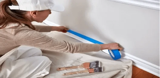 Woman in hat and overalls using painters&rsquo; tape to prep wall for painting.